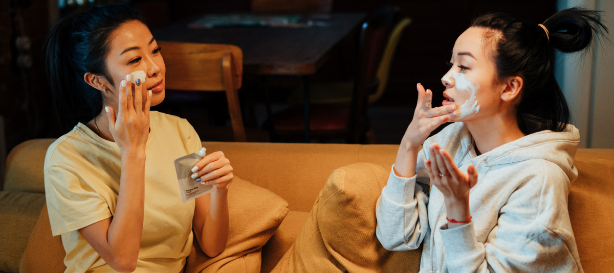 Two women sit on a couch and soothe themselves during the pandemic crisis by taking some time to apply a face mask, chat and relax.