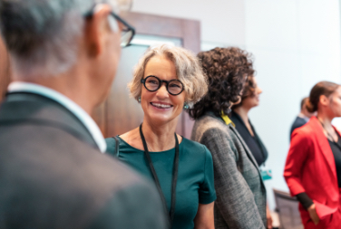 Two people wearing name tags around their necks smile at each other with other event attendees in the background