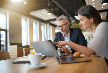 A man and a woman sit at a table in an office while the woman points at something on a laptop screen