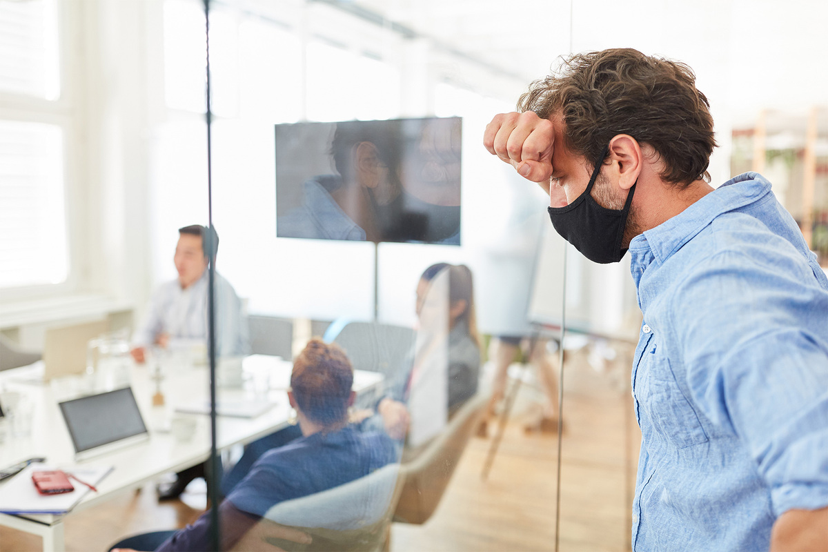 Un employé souffrant d'anxiété et portant un masque se trouve à l'extérieur d'une salle de réunion en essayant de retrouver son calme pendant que ses collègues se rassemblent à l'intérieur. Nos conseils d'experts peuvent aider quiconque à surmonter l'anxiété.