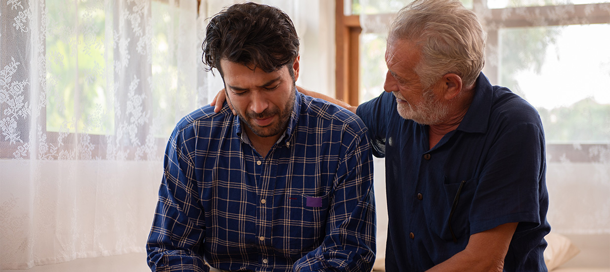 Two men sit next to each other in a living room discussing men and suicide. The older man (right) comforts the younger man (left), by putting an arm around him.