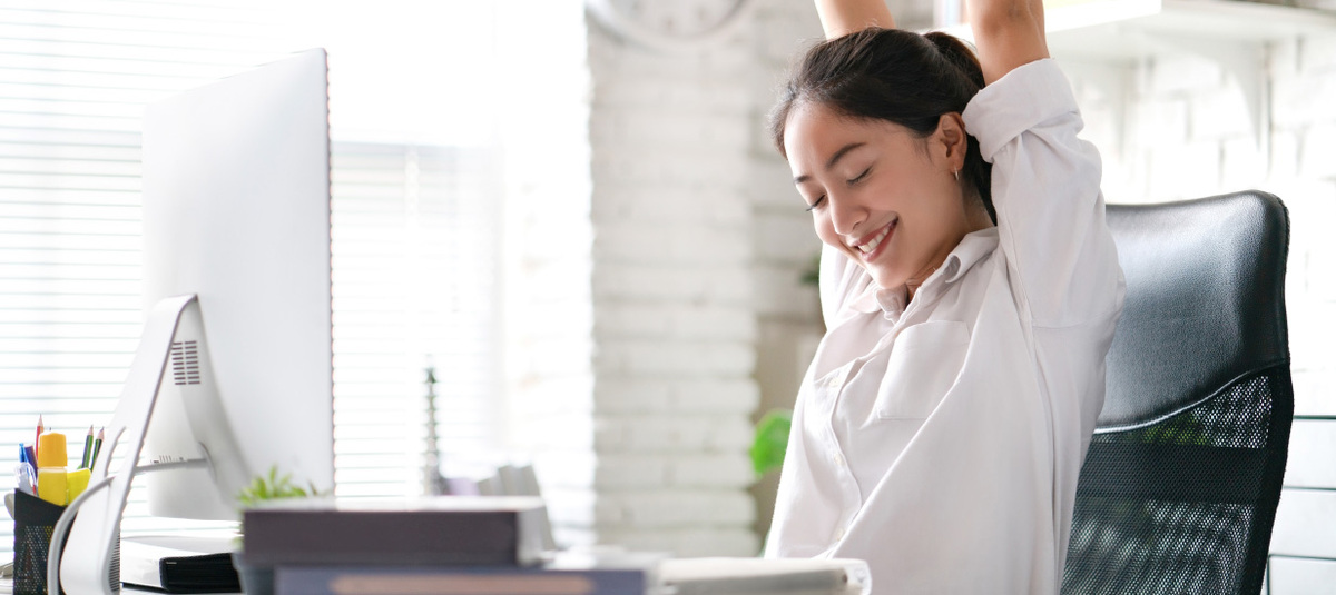 A woman stretches at her arms above her head and smiles at her ergonomic workstation.