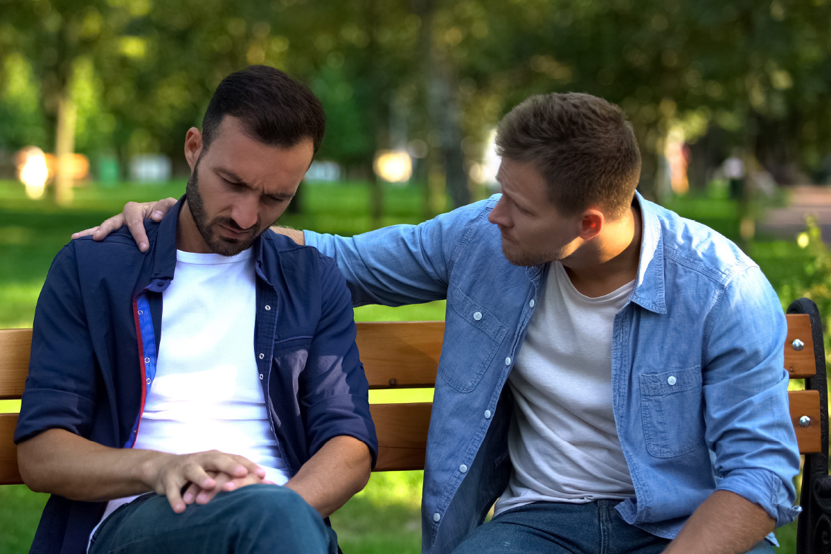 Deux hommes assis l'un à côté de l'autre dans un salon. Le plus âgé (à droite) réconforte le plus jeune bouleversé (à gauche) en plaçant son bras autour de lui.