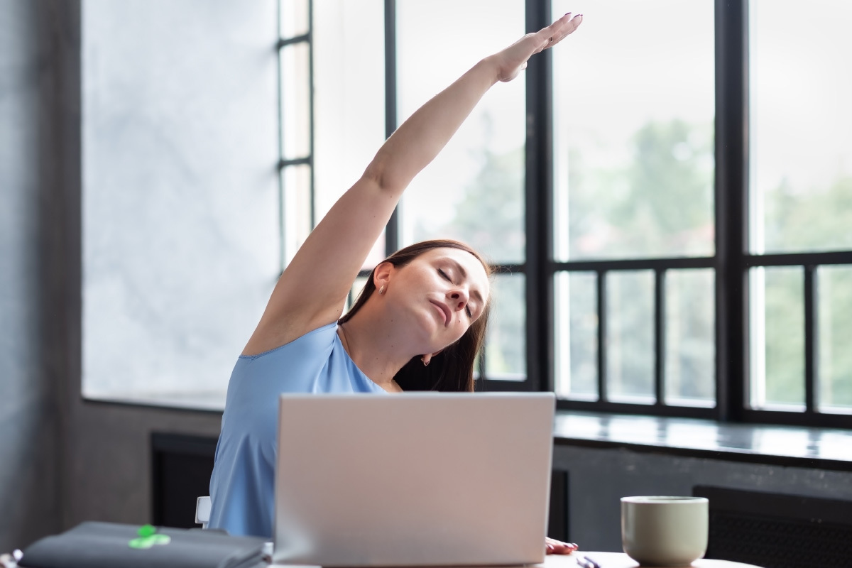A woman stretches at her desk to exercise while working from home.