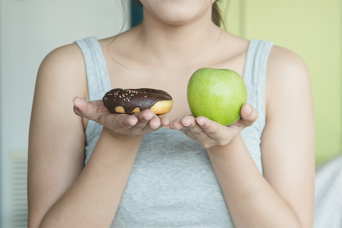 A woman holds an apple in one hand and a donut in the other, trying to make the decision to build better habits.