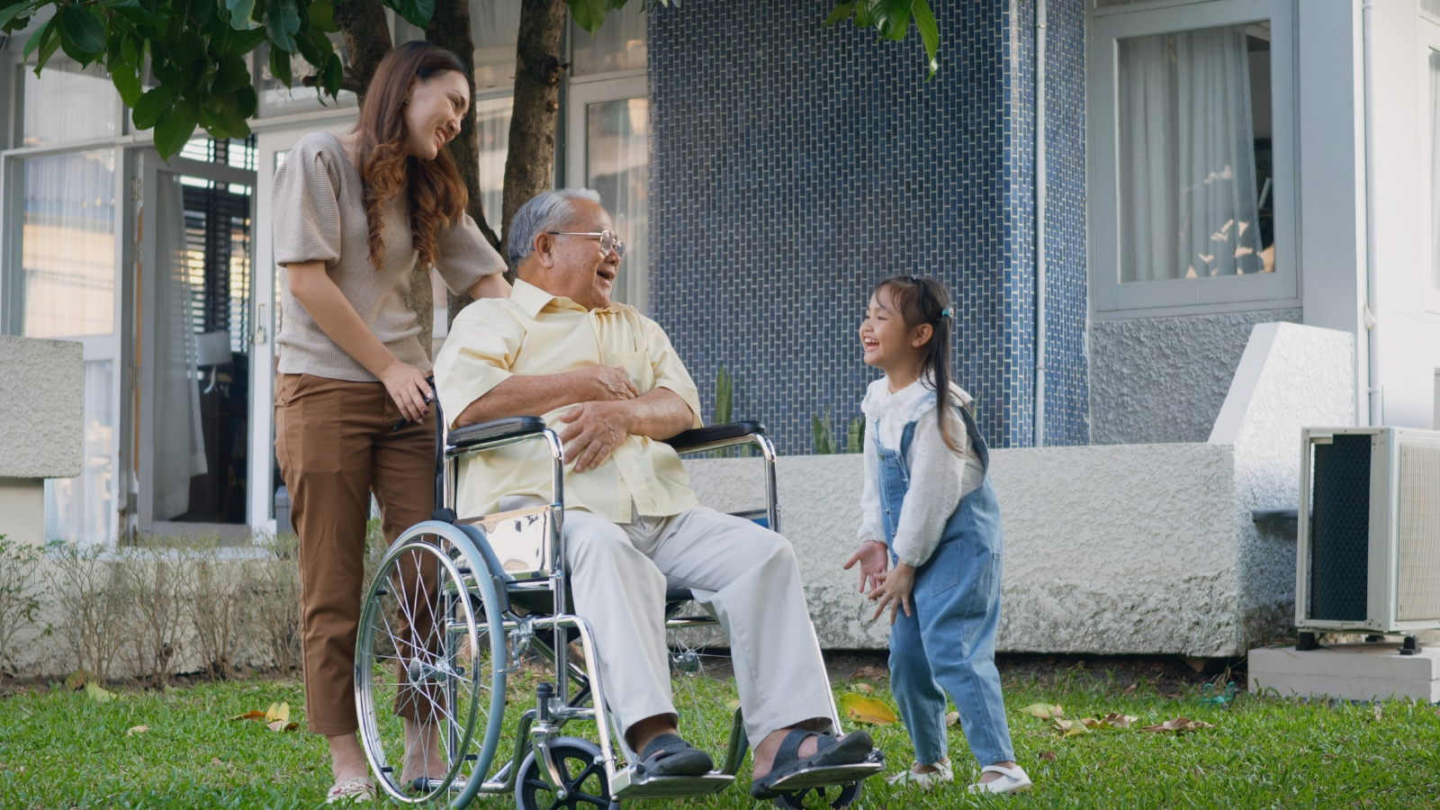 A mother and daughter share caregiving responsibilities by taking a grandparent in a wheelchair out for a walk.