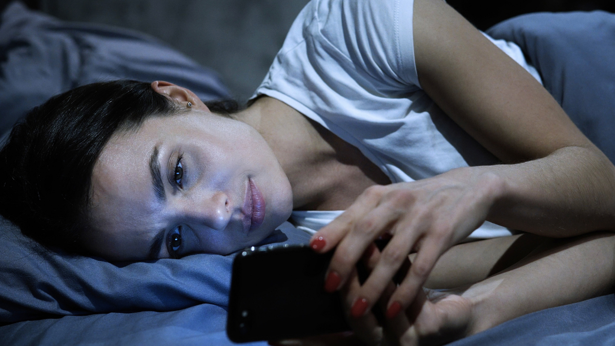 A woman lies in bed at night staring at social media on her phone.