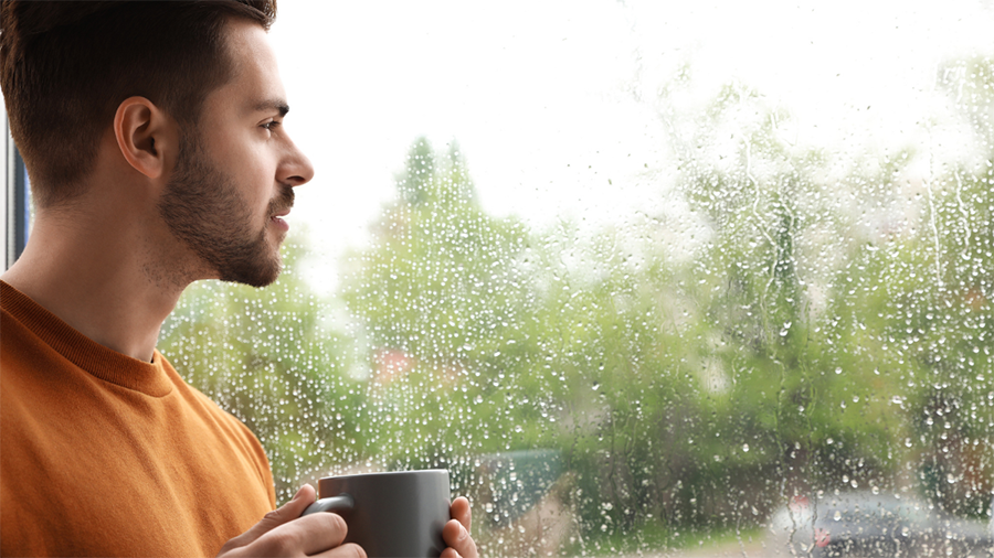 A man drinks coffee and gazes out the window during a rainy day, contemplating eco worry.