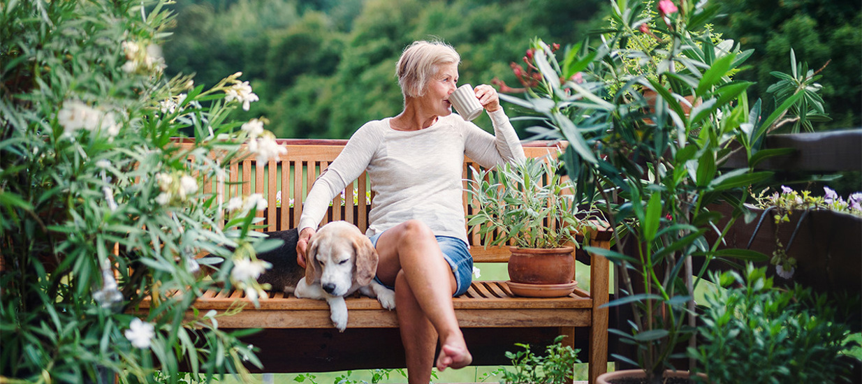 Woman sitting on a bench in a garden, sipping tea with a dog next to her