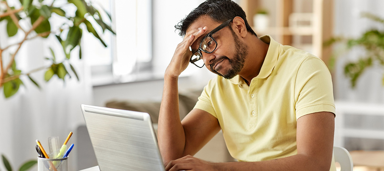 A man sitting at a computer with his right hand above his brow, looking stressed