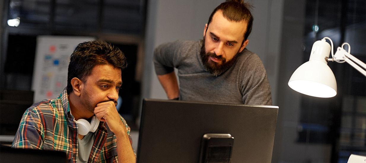 A manager looks over the shoulder of an employee sitting at a desk late at night