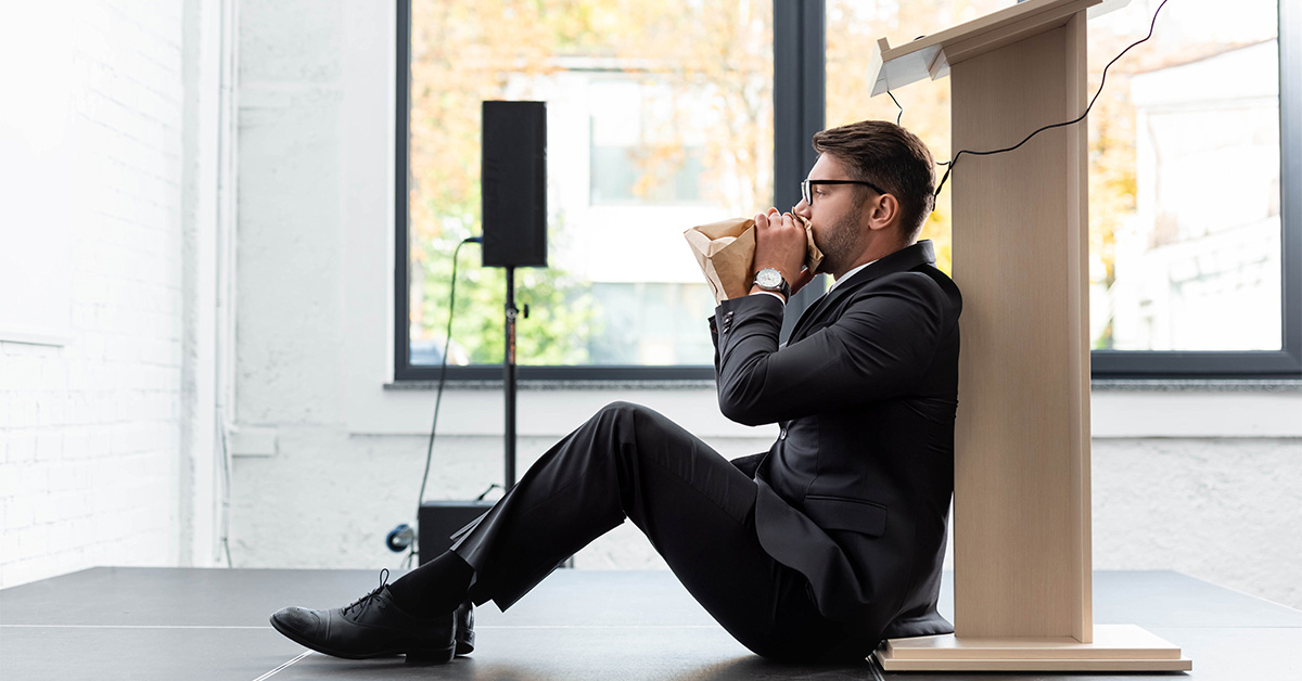 Man in a suit sitting on the ground behind a podium, breathing into a paper bag