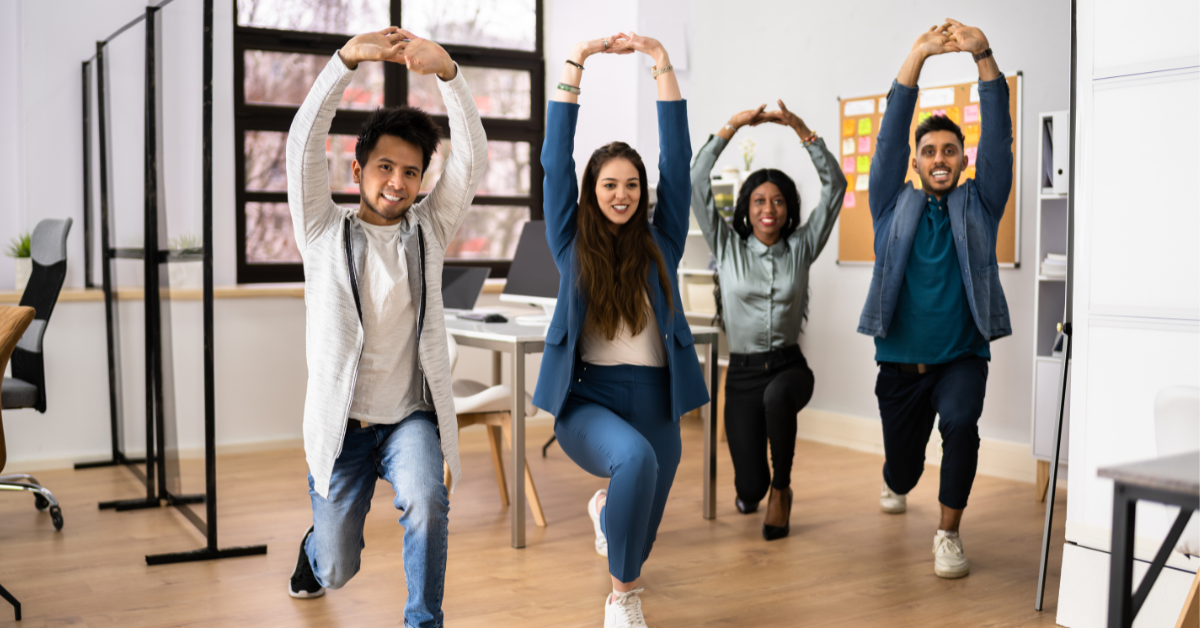 A group of four employees lunging forward in an office. They are participating in a workplace stretching session.