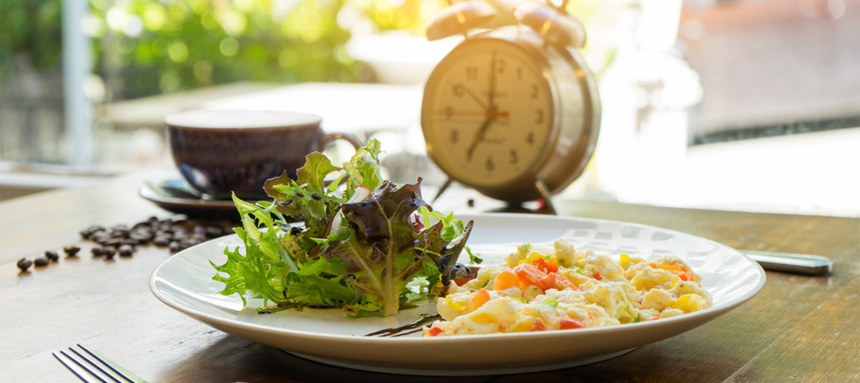 A plate of eggs and salad on a wooden table in the foreground. A windup clock indicating 7 o'clock in the background beside a cup of coffee on a saucer. There are coffee beans next to the coffee cup.