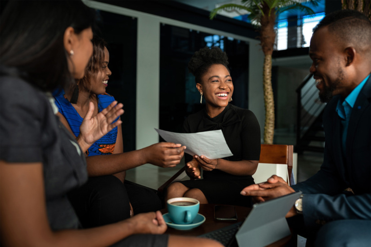 A group of black employees sit in a meeting. A cup of coffee is on the table in front of a woman holding a document.