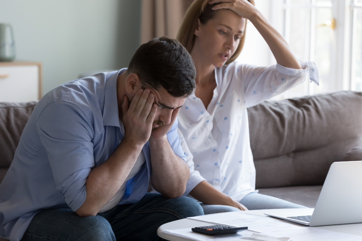 A man and a woman are sitting on a couch staring at a computer, with a calculator next to it. The woman has one of her hands on her head in disbelief. The man has his head in his hands, looking stressed.