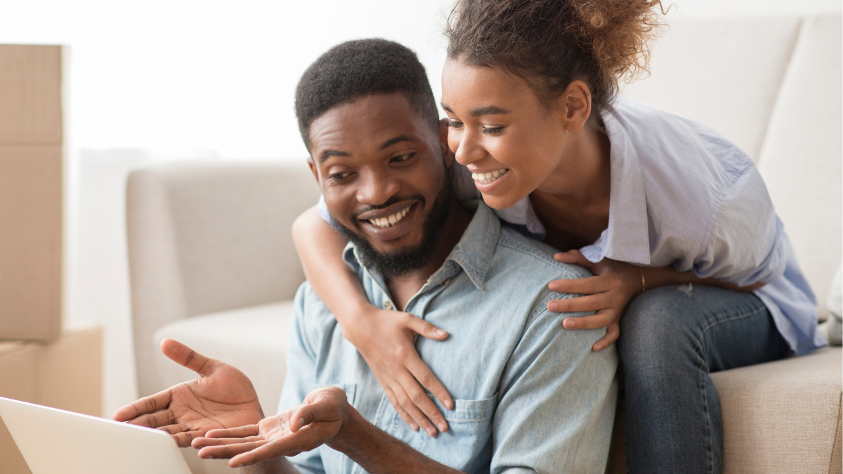 A woman sitting on a couch, leaning forward on her male partner who's showing her something on his laptop.