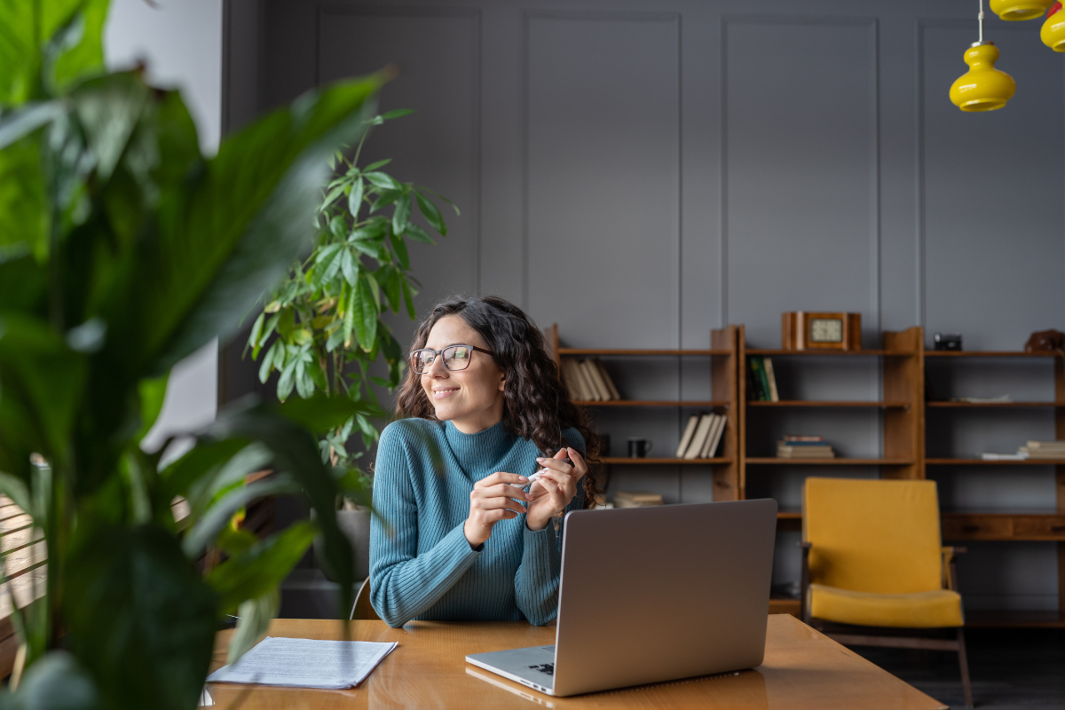 A woman in a green sweatshirt sits at a desk with a laptop open in front of her. She is looking out the window. There are green plants to the right of her and a bookshelf behind her. She is smiling.