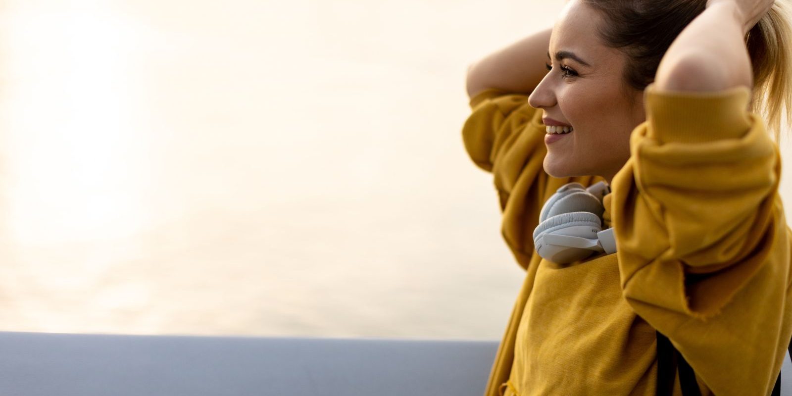 Smiling woman in yellow hoodie by the water, adjusting hair, headphones around neck.