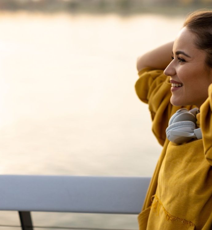 Smiling woman in yellow hoodie by the water, adjusting hair, headphones around neck.