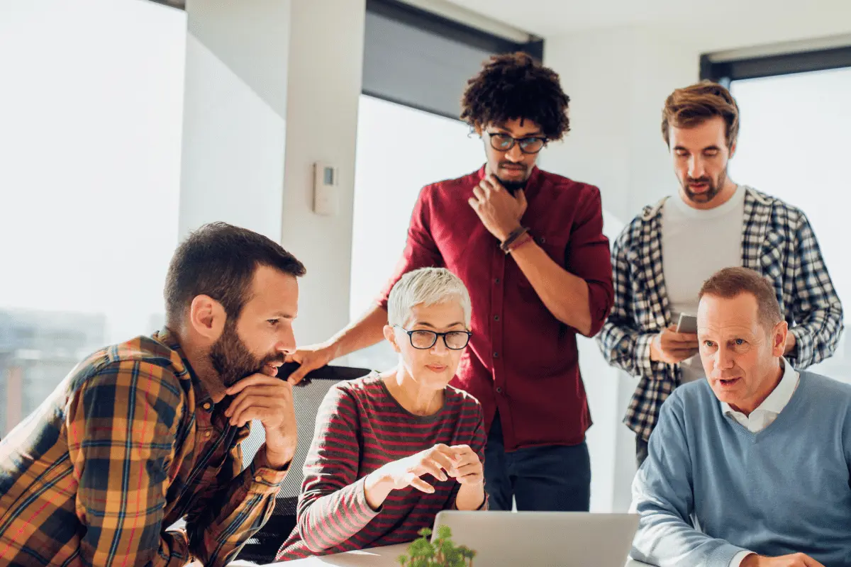 A diverse group of professionals gathered around a laptop in a bright office, collaborating on workplace strategies for supporting employee wellness goals.