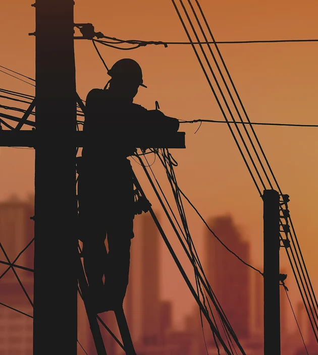 Silhouette of a worker on a utility pole with cityscape at sunset.