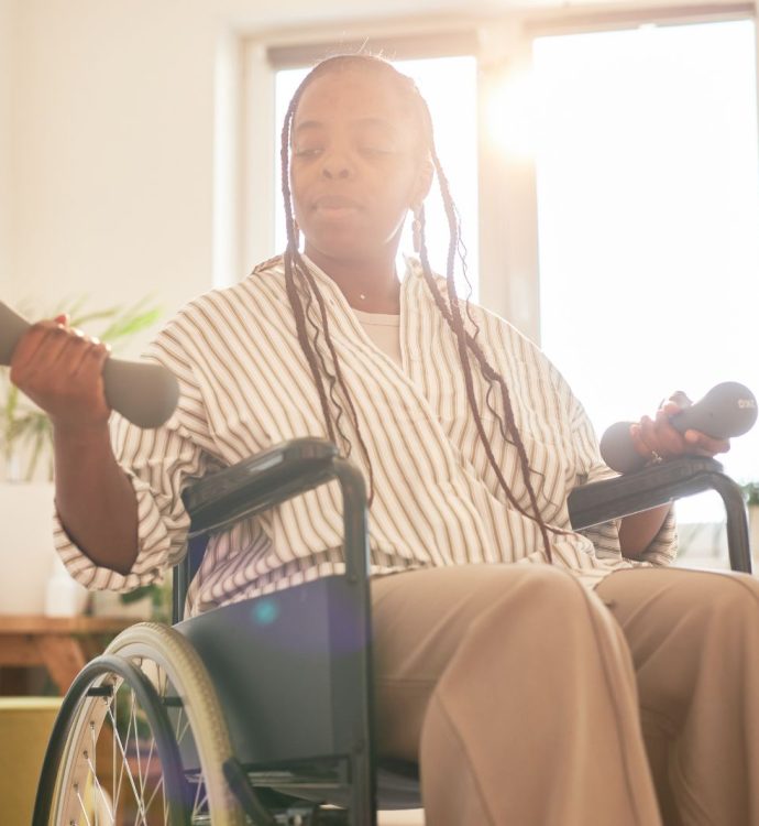 Person in a wheelchair lifting dumbbells in a sunlit room for exercise and well-being.