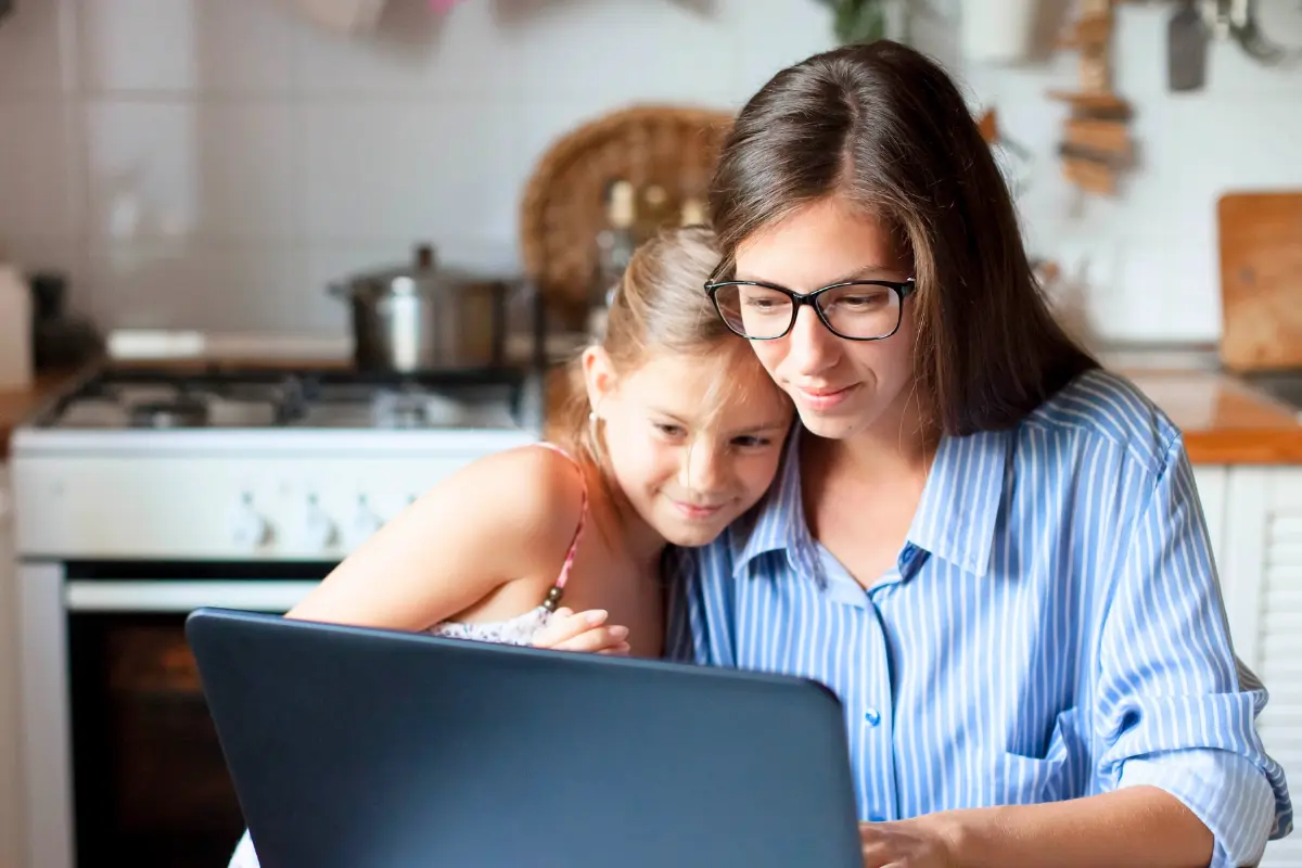 A parent and child sitting together in a kitchen, looking at a laptop, illustrating the challenges of balancing work and caregiving responsibilities during summer.