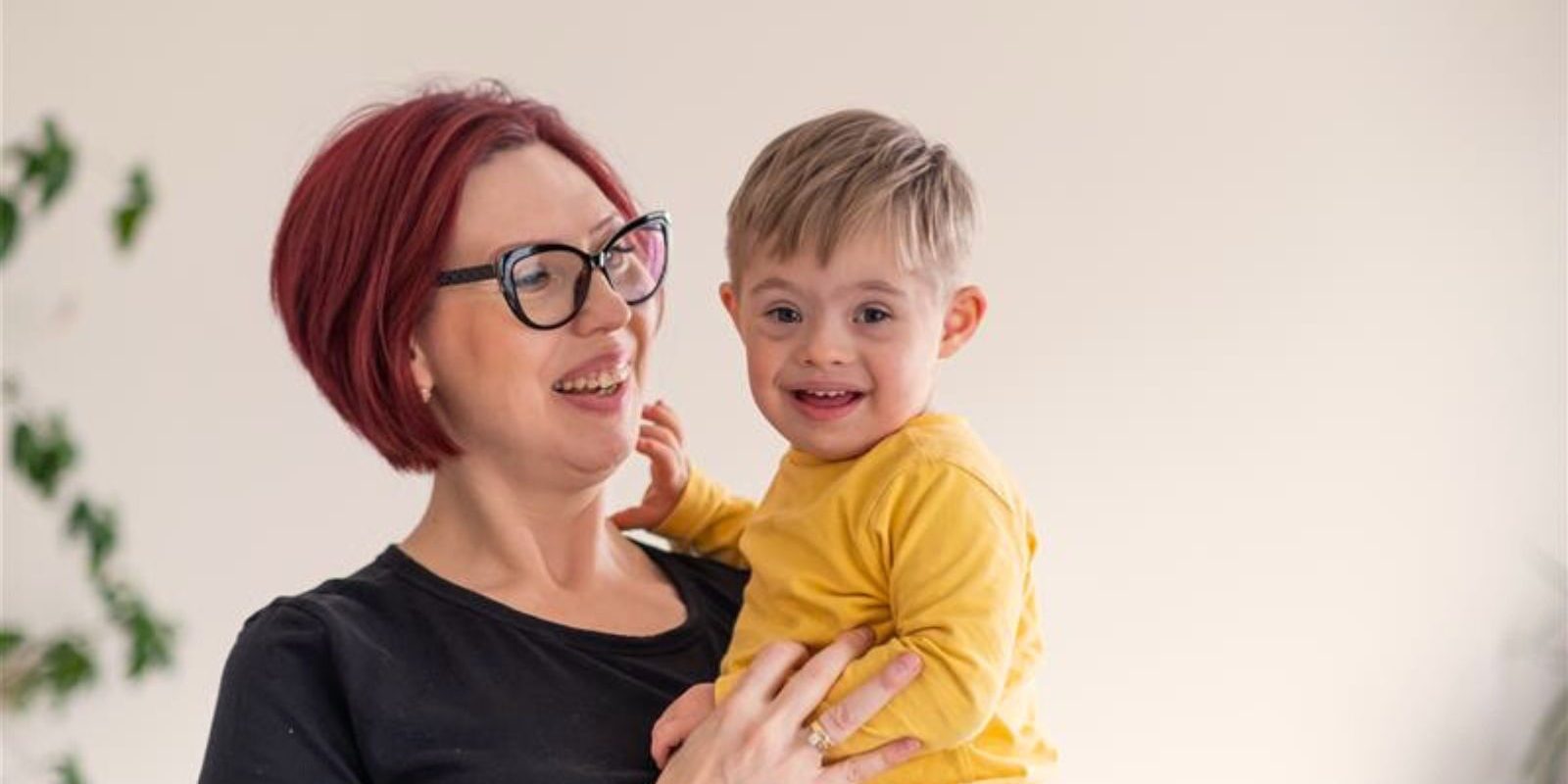 Smiling woman holding happy child in yellow shirt, indoor setting with plants in background.