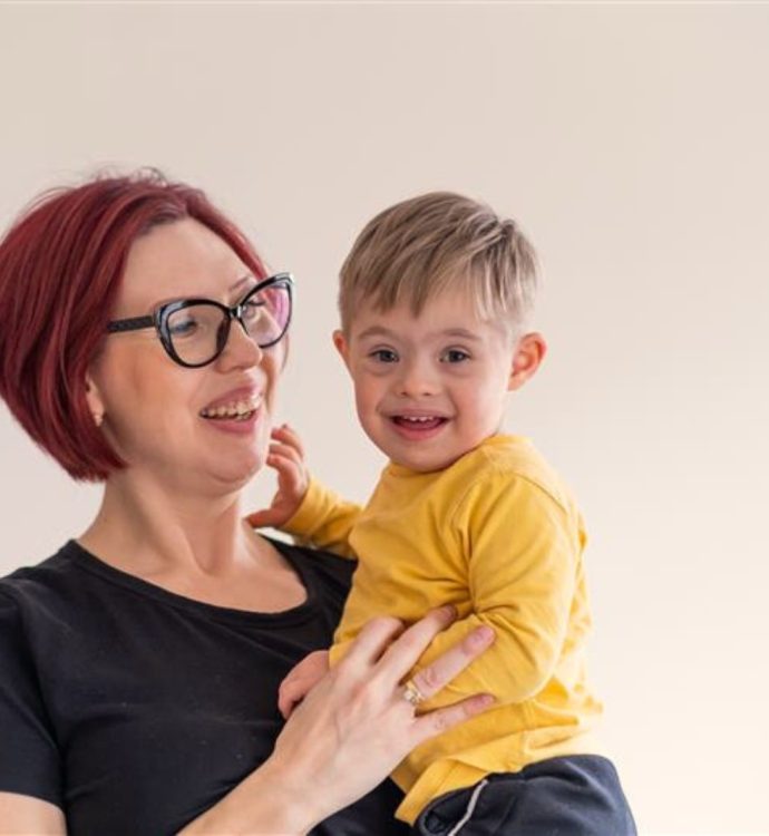 Smiling woman holding happy child in yellow shirt, indoor setting with plants in background.