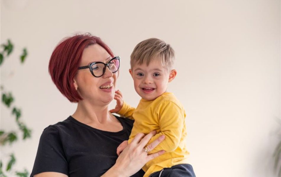 Smiling woman holding happy child in yellow shirt, indoor setting with plants in background.