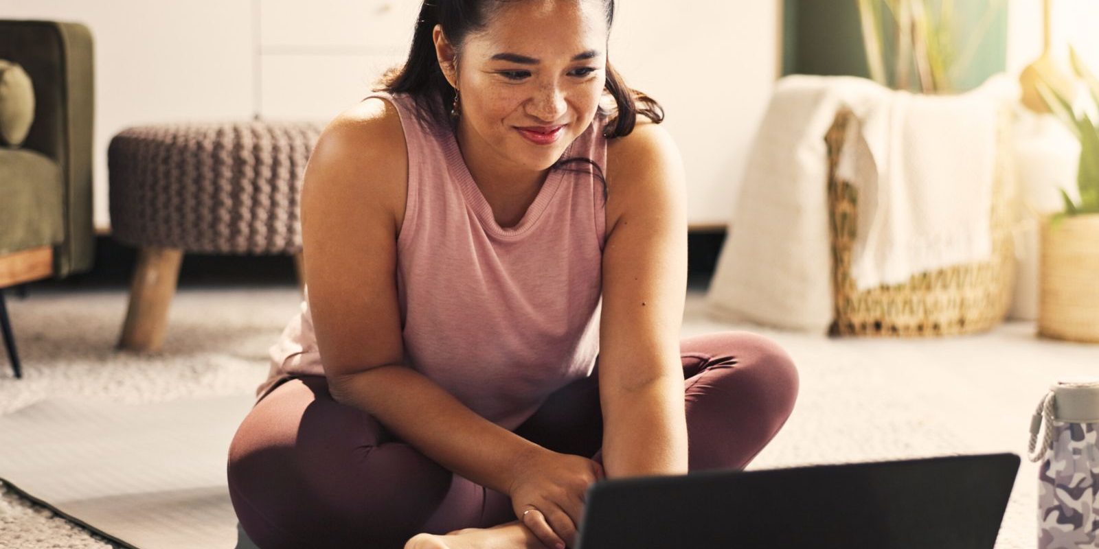 Woman engaged in online workout session at home on yoga mat, smiling and focused on laptop.
