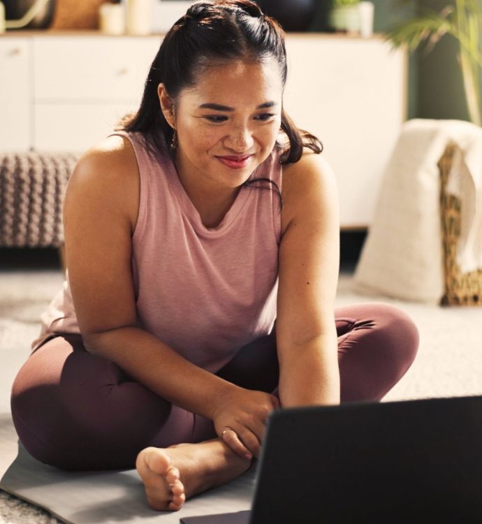 Woman engaged in online workout session at home on yoga mat, smiling and focused on laptop.