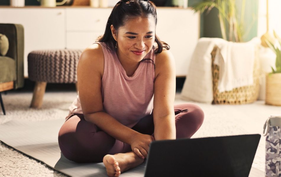 Woman engaged in online workout session at home on yoga mat, smiling and focused on laptop.