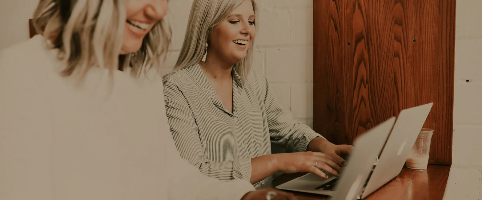 Women smiling while working on laptops at a wooden table.