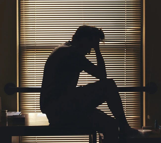 Silhouette of a person sitting by a window, looking stressed with hand on head.