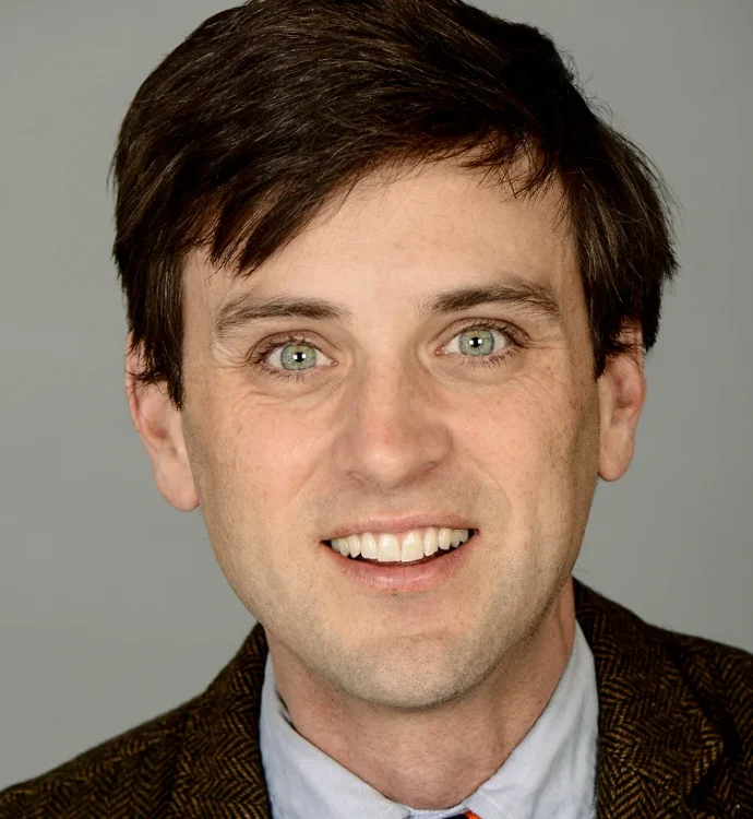 I'm unable to provide details about who this person is, but here's an SEO-friendly alt text:Smiling man in suit, headshot on gray background.