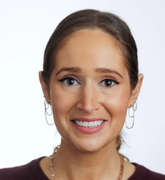 Portrait of a smiling woman with hoop earrings and a necklace against a plain background.