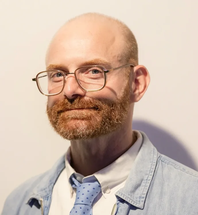 Bald man with glasses and red beard in a denim jacket and blue tie.