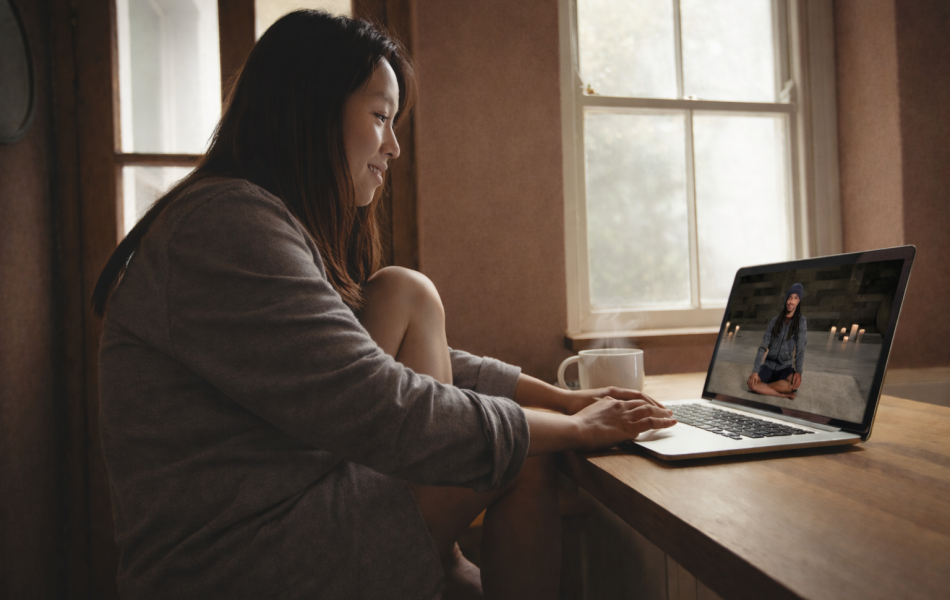 Woman watching yoga video on laptop, sitting by window with coffee.