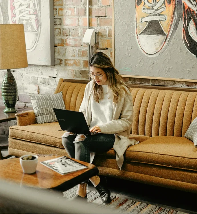 Woman working on laptop in cozy, vintage living room with art on brick walls.