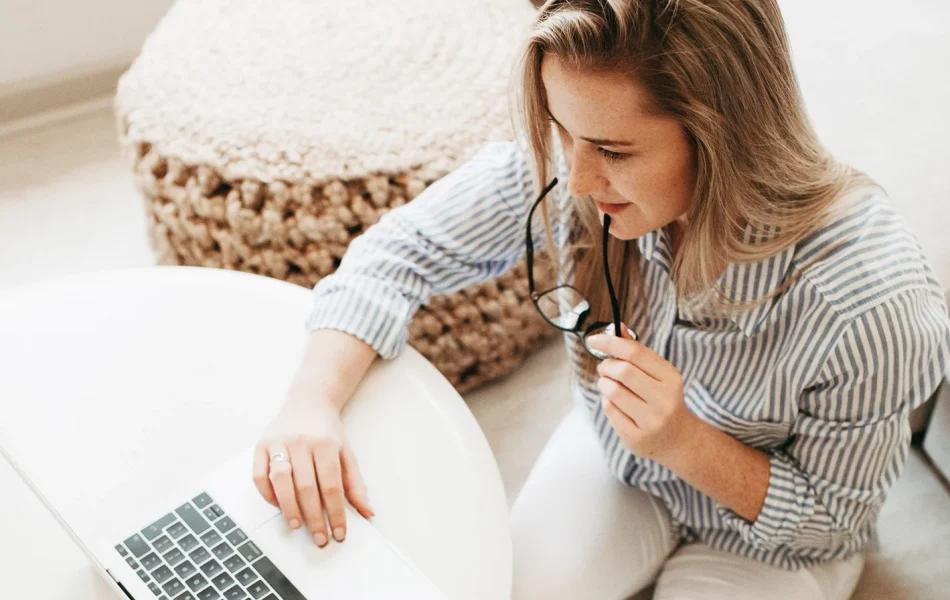 Woman working on laptop at white table, holding glasses thoughtfully.