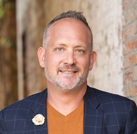 Smiling man in blazer and orange shirt, professional headshot against textured wall