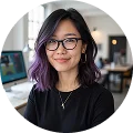 Smiling woman with glasses and purple hair at modern office desk, computer in background.