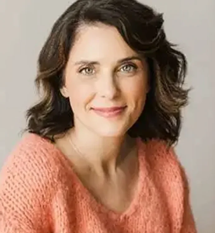 Smiling woman in a cozy pink sweater with wavy brown hair against a neutral background.