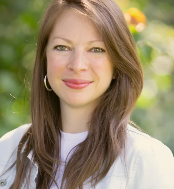 Smiling woman in garden, wearing hoop earrings and white jacket.