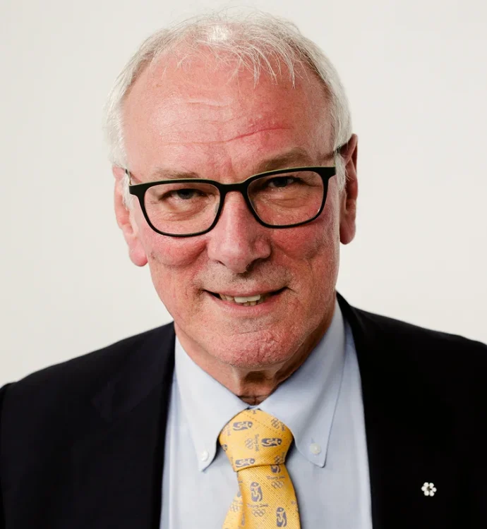 Elderly man in suit and glasses smiling against a plain background.