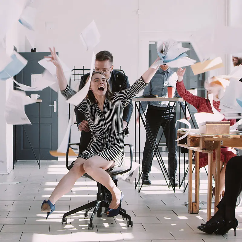 Office celebration with woman in chair, papers flying, colleagues cheering, bright workspace.
