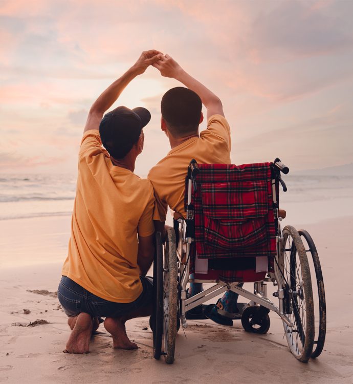 Two people on a beach at sunset, one in a wheelchair, forming a heart with their hands.