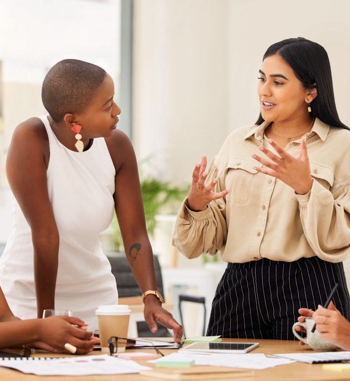Women engaging in a lively office discussion around a table with documents and coffee.